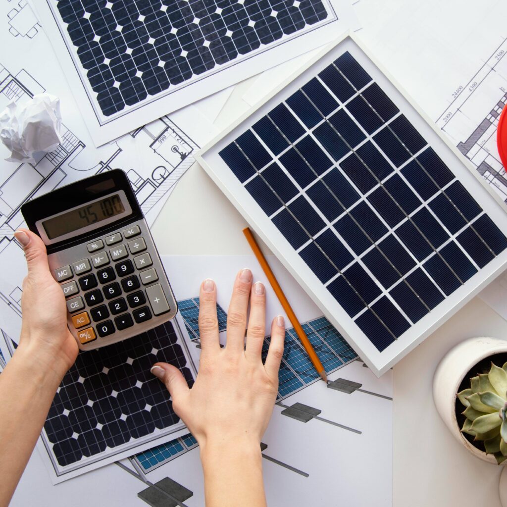Person calculating solar panel system design with blueprints and miniature solar panels on desk